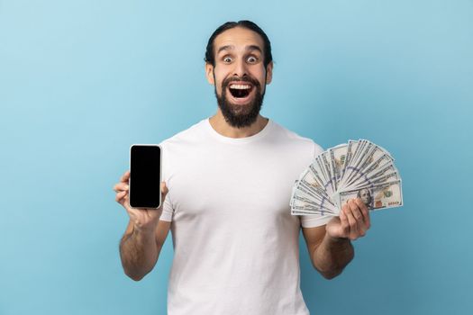 Portrait of man with beard wearing white T-shirt holding fan of dollar banknotes and smart phone with black empty display, making money online. Indoor studio shot isolated on blue background.
