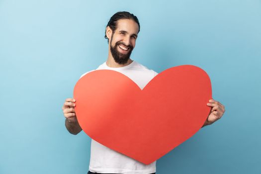 Portrait of romantic man with beard wearing white T-shirt holding in hands big red paper heart and smiling at camera, love and romance. Indoor studio shot isolated on blue background.