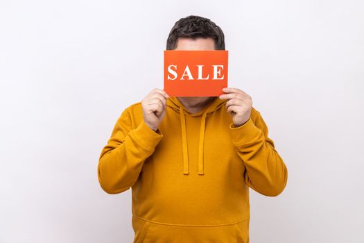 Portrait of unknown anonymous man covering eyes with Sale inscription, market discounts, black friday, wearing urban style hoodie. Indoor studio shot isolated on white background.
