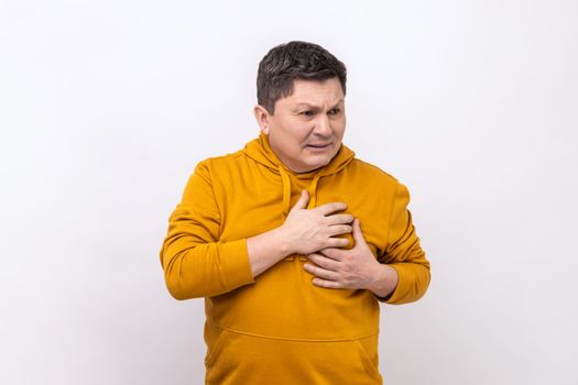 Heart attack or problem. Portrait of sad shocked handsome man standing and holding his chest with painful face, wearing urban style hoodie. Indoor studio shot isolated on white background.