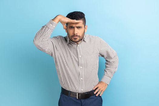 Portrait of handsome businessman looking into distance, holding hand over eyes and watching horizon with attentive view, wearing striped shirt. Indoor studio shot isolated on blue background.