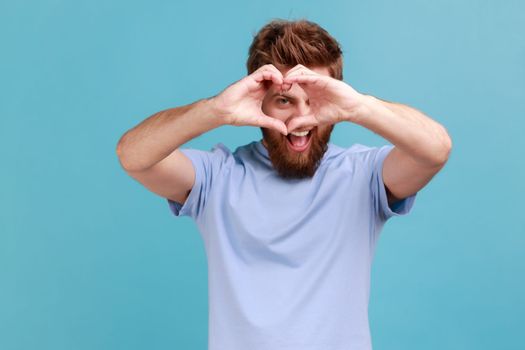 Portrait of handsome bearded man looking through hands in shape of heart showing romantic gesture, love confession, valentines day celebration. Indoor studio shot isolated on blue background.