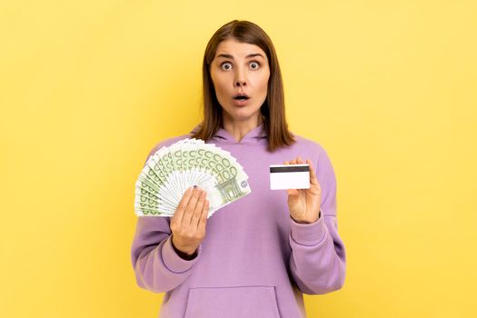 Portrait of shocked woman with dark hair holding fan of euro banknotes and credit card, earning money, banking, wearing purple hoodie. Indoor studio shot isolated on yellow background.