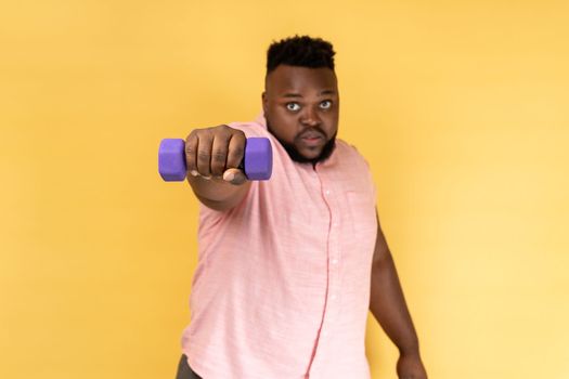 Portrait of serious self confident bearded man wearing pink shirt training his arms with dumbbell, looking at camera, healthy lifestyle. Indoor studio shot isolated on yellow background.