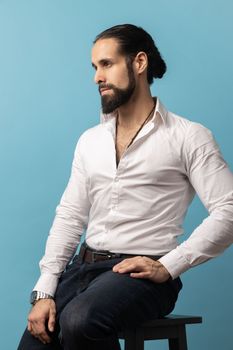 Side view of handsome man with beard and dark collected hair wearing white shirt and black trousers, sitting and looking away with pensive expression. Indoor studio shot isolated on blue background