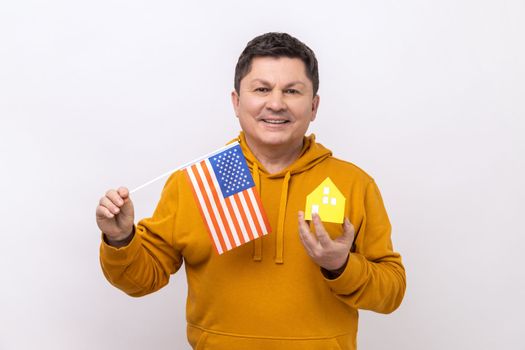 Portrait of middle aged man holding american flag and paper house, dreaming to buy accommodation in USA, wearing urban style hoodie. Indoor studio shot isolated on white background.
