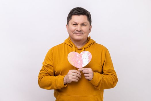 Playful middle aged man holding heart sticks and smiling, holding pink greeting card in heart shape, preparing gift, wearing urban style hoodie. Indoor studio shot isolated on white background.