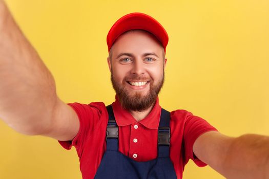 Optimistic positive worker man wearing blue uniform taking selfie, looking at camera with toothy POV, point of view of photo. Indoor studio shot isolated on yellow background.