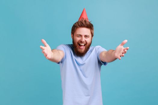 Portrait of extremely happy bearded man wearing party cone giving free hugs with outstretched hands, welcoming inviting to embrace. Indoor studio shot isolated on blue background.
