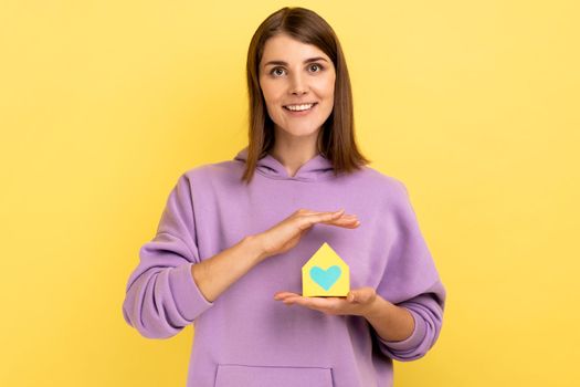 Portrait of woman holding in hands near heart little paper house looks at camera with pleasant smile, dreaming about own house, wearing purple hoodie. Indoor studio shot isolated on yellow background.