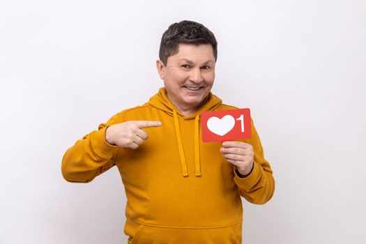 Internet blogging. Portrait of joyful man pointing at heart like icon, recommending to click on social media button, wearing urban style hoodie. Indoor studio shot isolated on white background.