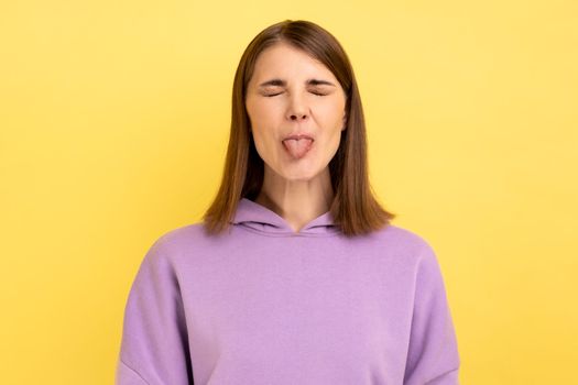 Portrait of funny disobedient young woman demonstrating tongue, behaving naughty unruly, childish mood, closed eyes, wearing purple hoodie. Indoor studio shot isolated on yellow background.