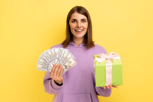 Happy optimistic woman holding wrapped present box and dollars banknotes, looking at camera with positive expression, wearing purple hoodie. Indoor studio shot isolated on yellow background.