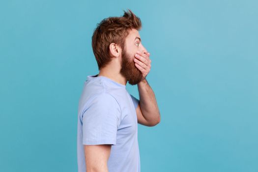 I don't tell anyone. Side view of handsome young adult bearded man zipping mouth, keeping secret information, looking at side space. Indoor studio shot isolated on blue background.