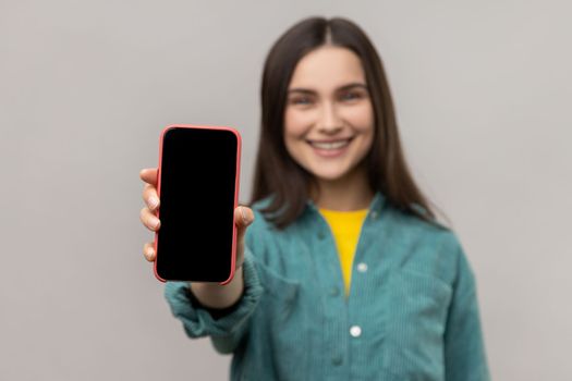 Portrait of smiling friendly woman showing cell phone with empty display with copy space for advertisement, wearing casual style jacket. Indoor studio shot isolated on gray background.