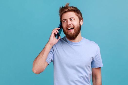 Portrait of excited handsome bearded man standing, talking smartphone, making call and having pleasant conversation with friend or wife. Indoor studio shot isolated on blue background.