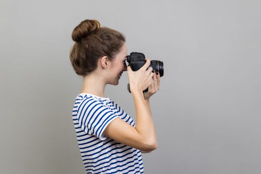 Side view of woman photographer or traveler wearing striped T-shirt holding professional digital dslr camera and focusing, taking photo, shooting video. Indoor studio shot isolated on gray background.