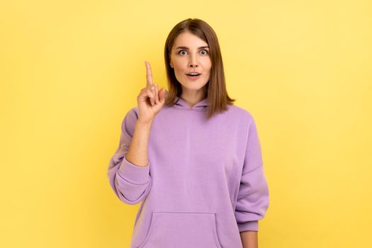 Portrait of inspired woman raising finger and having genius idea, looking amazed by sudden smart thought, wearing purple hoodie. Indoor studio shot isolated on yellow background.