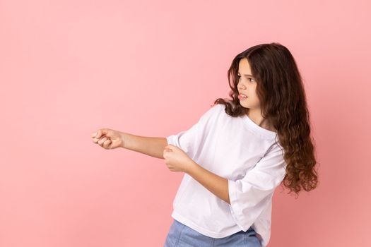 Side view portrait of adorable charming little girl wearing white T-shirt standing in attack or pulling hands gesture and looking forward. Indoor studio shot isolated on pink background.