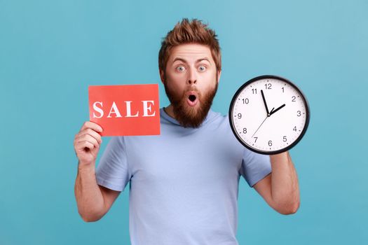Portrait of shocked astonished handsome bearded man holding clock and card with sale inscription, announcing finishing huge discounts. Indoor studio shot isolated on blue background.