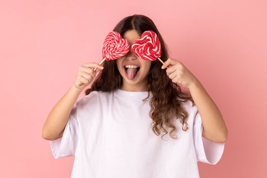 Portrait of little girl wearing white T-shirt covering her eyes with heart shape candies and showing tongue out, posing with happy expression. Indoor studio shot isolated on pink background.