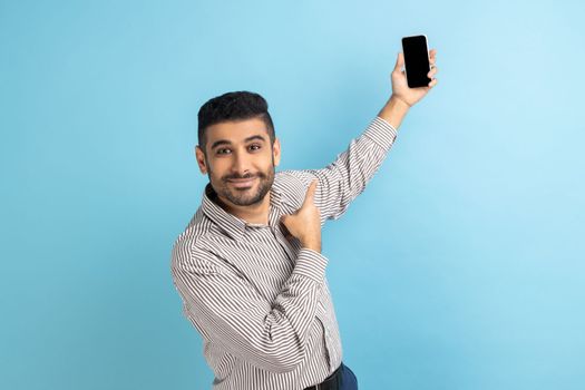 Portrait of businessman talking video call, looking at camera, pointing at smartphone up with mockup blank display, wearing striped shirt. Indoor studio shot isolated on blue background.