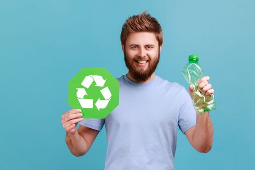 Portrait of smiling optimistic handsome bearded man pressing bottle holding in hand recycling sign board, plastic disposal, garbage sorting. Indoor studio shot isolated on blue background.