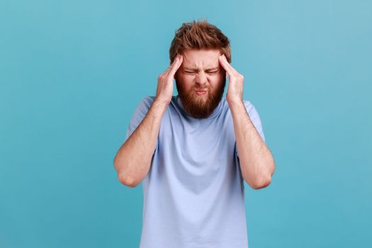 Portrait of unhealthy bearded man massaging temples feeling headache, suffering migraine or high blood pressure, trying to concentrate. Indoor studio shot isolated on blue background.