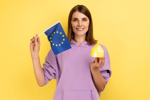 Real estate purchase in EU, immigration. Portrait of satisfied woman holding European flag and paper house, wearing purple hoodie. Indoor studio shot isolated on yellow background.