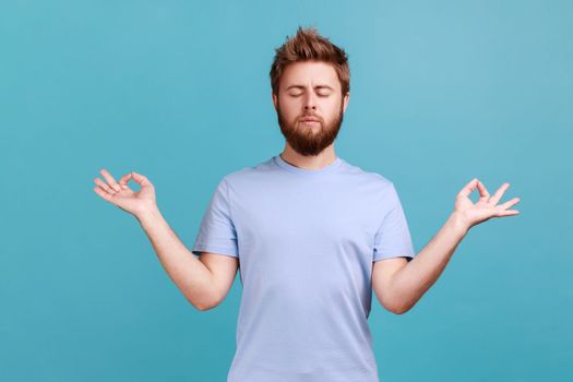 Portrait of calm man standing with raised arms and doing yoga meditating exercise, keeping eyes closed, practicing yoga mudras to relax and concentrate. Indoor studio shot isolated on blue background.