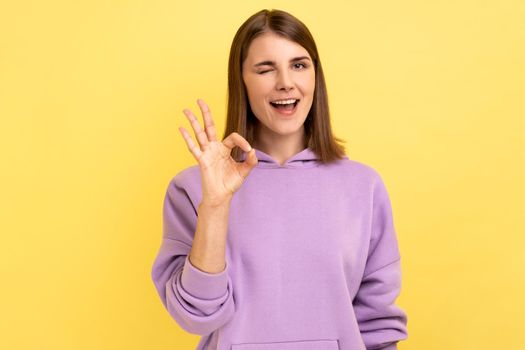 Woman with toothy smile, winking to camera and showing thumb up, approval sign, satisfied with service, good feedback, wearing purple hoodie. Indoor studio shot isolated on yellow background.