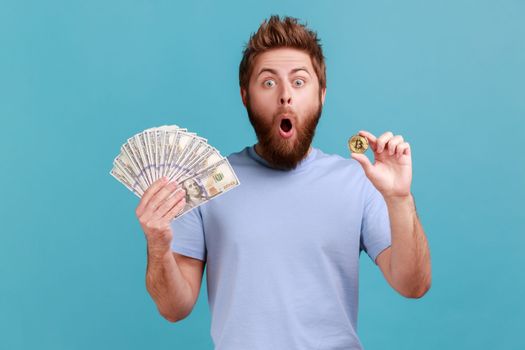 Portrait of amazed surprised handsome bearded man showing dollar banknotes and bitcoin, digital money, electronic commerce, expressing astonishment. Indoor studio shot isolated on blue background.