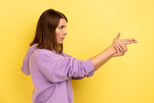 I'll kill you. Side view of angry woman pointing finger guns to camera, threatening to shoot, hands imitating weapon, wearing purple hoodie. Indoor studio shot isolated on yellow background.