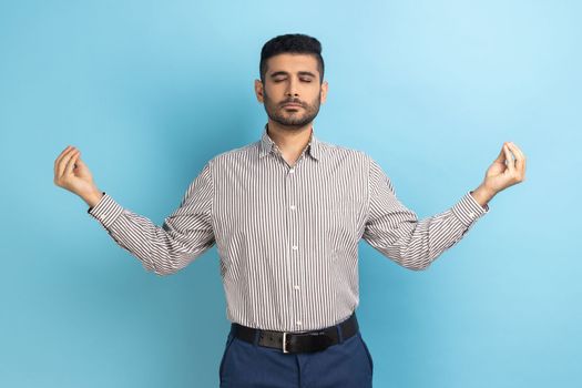 Portrait of calm relaxed young businessman with beard standing with raised arms and doing yoga meditating exercise, wearing striped shirt. Indoor studio shot isolated on blue background.