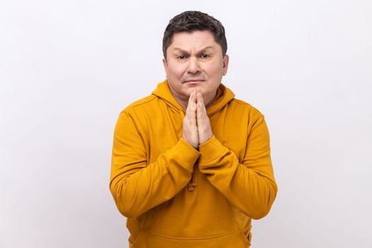 Please, I'm begging. Portrait of upset brunette man holding arms in prayer, asking help or forgiveness with imploring eyes, wearing urban style hoodie. Indoor studio shot isolated on white background.