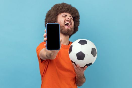 Extremely happy man with Afro hairstyle wearing T-shirt standing with soccer ball and showing mobile phone with empty display, betting and winning. Indoor studio shot isolated on blue background.