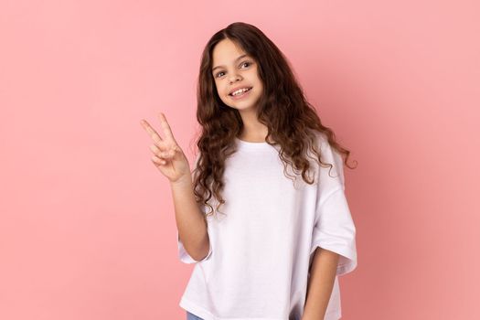 Portrait of positive optimistic little girl wearing white T-shirt looking at camera with toothy smile, showing v sign, peace gesture. Indoor studio shot isolated on pink background.