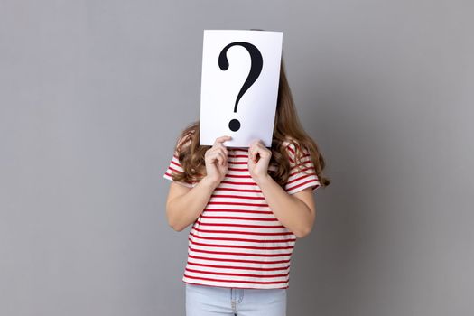 Portrait of unknown little girl wearing striped T-shirt standing hiding face behind paper with question mark, having problem for solving. Indoor studio shot isolated on gray background.