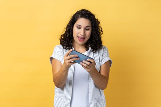 Leisure and technology. Woman using mobile phone with amazed expression, playing video game on cellphone, chatting in social network. Indoor studio shot isolated on yellow background.