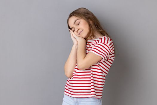 I need rest. Portrait of little girl wearing striped T-shirt yawning and covering mouth with hand, feeling exhausted, lack of sleep. Indoor studio shot isolated on gray background.
