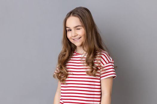 Portrait of playful cheerful little girl wearing striped T-shirt standing and winking playfully, positive expression, looking at camera. Indoor studio shot isolated on gray background.