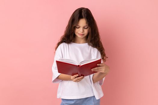 Portrait of delighted charming little girl wearing white T-shirt holding in hands and reading interesting book, being very attentive. Indoor studio shot isolated on pink background.