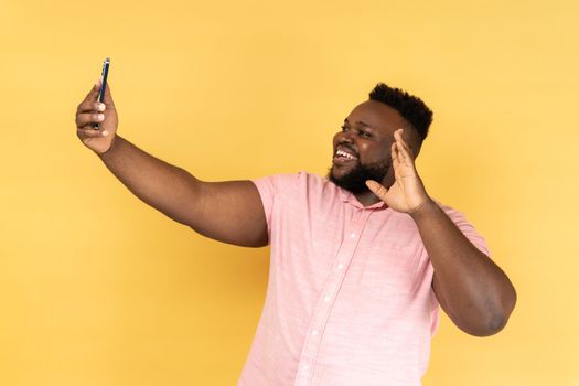 Portrait of bearded man wearing pink shirt saying hi making video call using smartphone, smiling with toothy smile, 4g connection. Indoor studio shot isolated on yellow background.