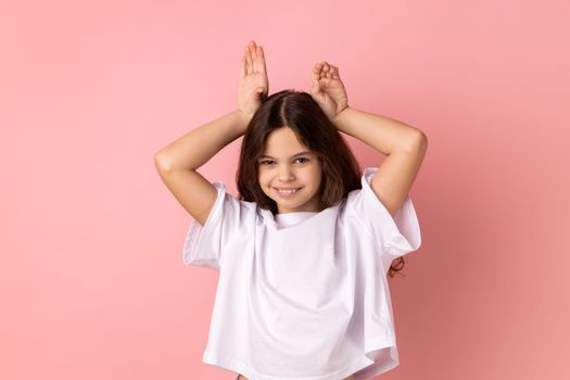 Portrait of positive little girl wearing white T-shirt making funny bunny ears with hands on head, childish behavior, playful optimistic mood. Indoor studio shot isolated on pink background.
