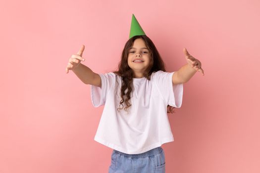 Portrait of delighted little girl wearing white T-shirt and party cone giving free hugs with outstretched hands, welcoming inviting to embrace. Indoor studio shot isolated on pink background.