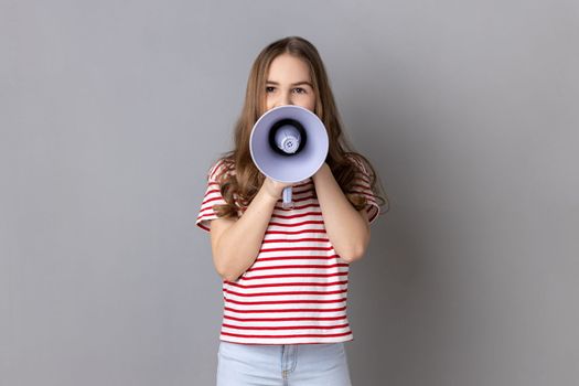 Portrait of dark haired little girl wearing striped T-shirt screaming in megaphone, speaking, announcing important information. Indoor studio shot isolated on gray background.