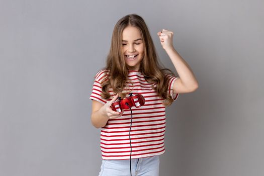 Portrait of little girl wearing striped T-shirt holding hand red joystick pad showing yes gesture sincerely rejoicing, victory in online virtual game. Indoor studio shot isolated on gray background.
