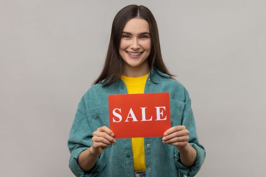 Portrait of young adult happy smiling woman standing holding card with sale inscription, huge discounts, wearing casual style jacket. Indoor studio shot isolated on gray background.
