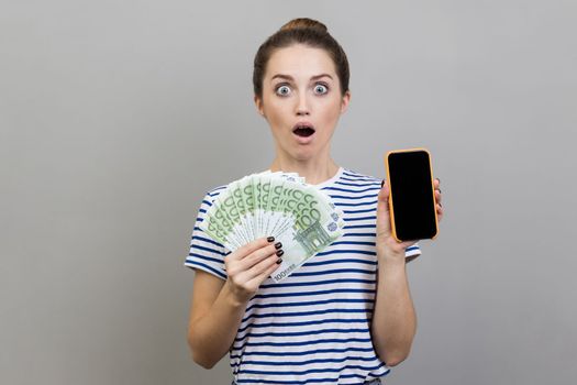 Portrait of shocked surprised young woman wearing striped T-shirt holding fan of euro banknotes and smart phone with black blank screen. Indoor studio shot isolated on gray background.