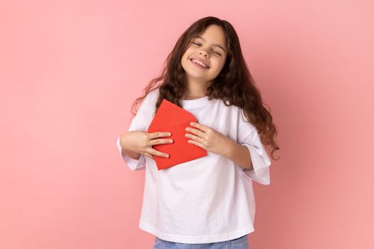 Portrait of satisfied little girl wearing white T-shirt standing open red envelope with congratulations, reading romantic letter, being touched. Indoor studio shot isolated on pink background.